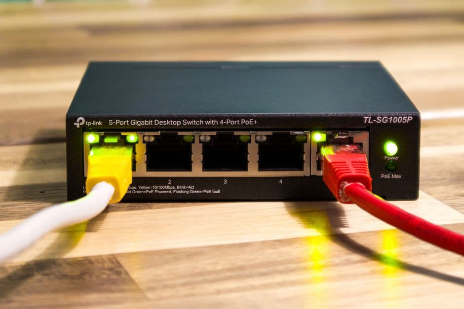 A black TP-Link TL-SG1005P five-port gigabit desktop switch on a wooden surface, with a white-and-yellow cable in port 1 and a red cable on the right, green link LEDs lit.