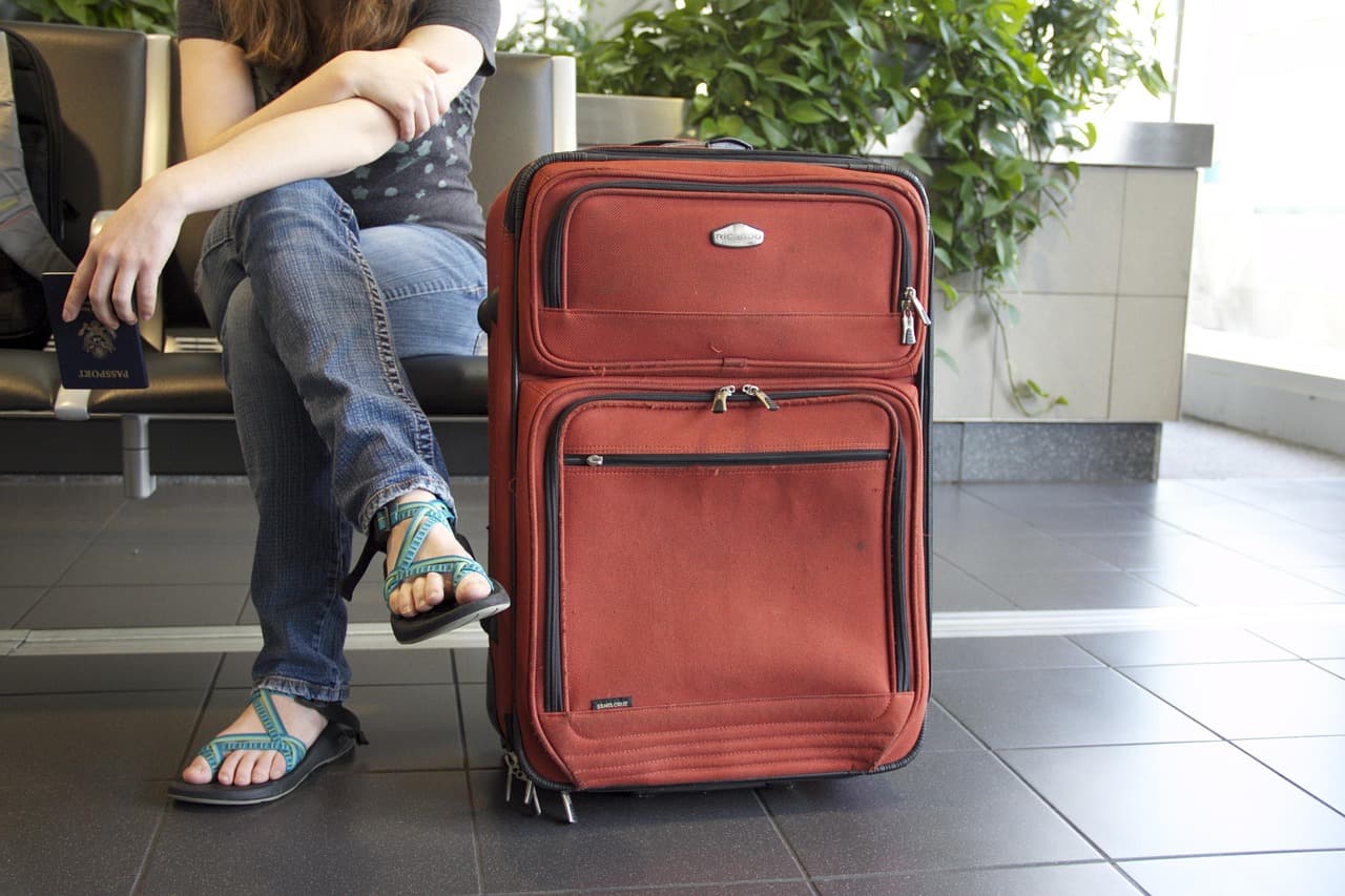 Une personne assise dans une salle d'attente d'aéroport tient un passeport, une valise à roulettes rouge posée à côté d'elle