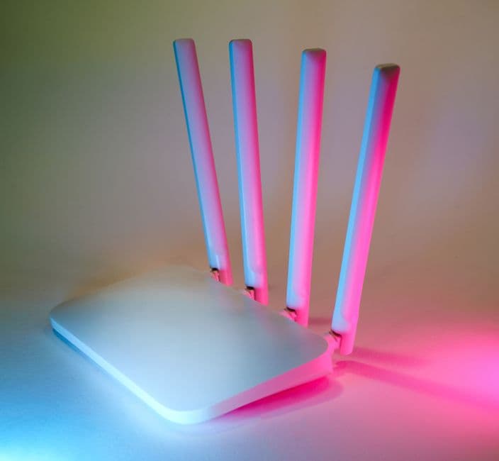 A white four-antenna wireless router photographed against a pale background, lit from the sides by pink and blue neon light