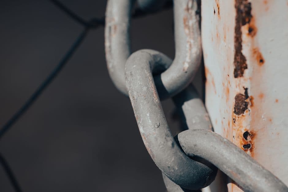 A close-up of a thick steel chain hanging against a white metal post streaked with rust, the background blurred.