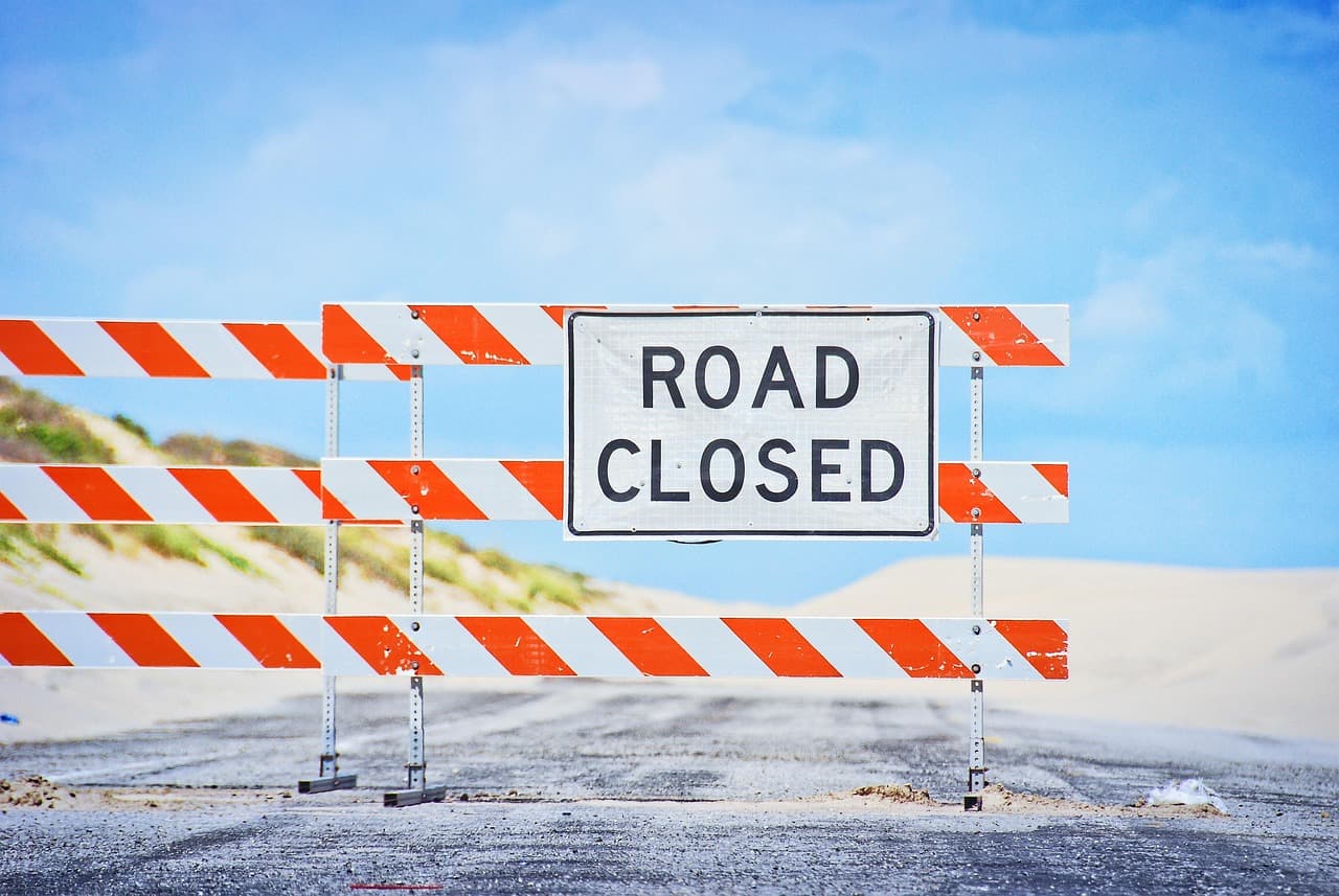 Eine Baustellenabsperrung mit dem Schild ROAD CLOSED vor einer sandbedeckten Straße