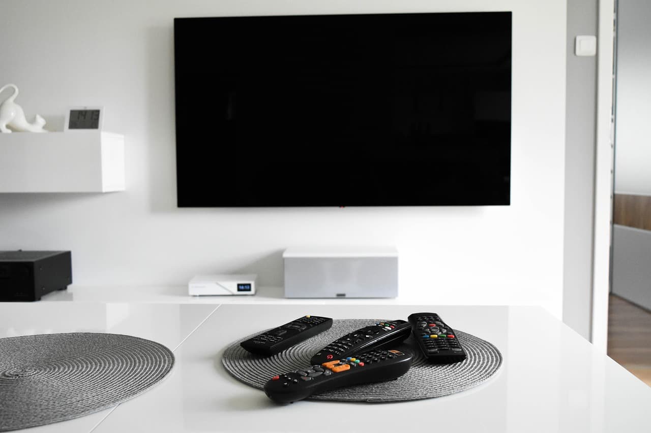 Um televisor de parede desligado, uma box na prateleira por baixo e quatro comandos pousados na mesa em frente.